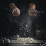 person pouring flour on table beside eggs and whisk