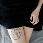 woman lying on bed sowing cat dreamcatcher tattoo on right leg during daytime